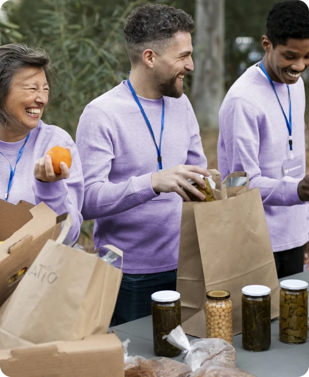 Grupo sorridente de voluntários vestindo suéteres roxos, organizando alimentos em sacolas para doação.