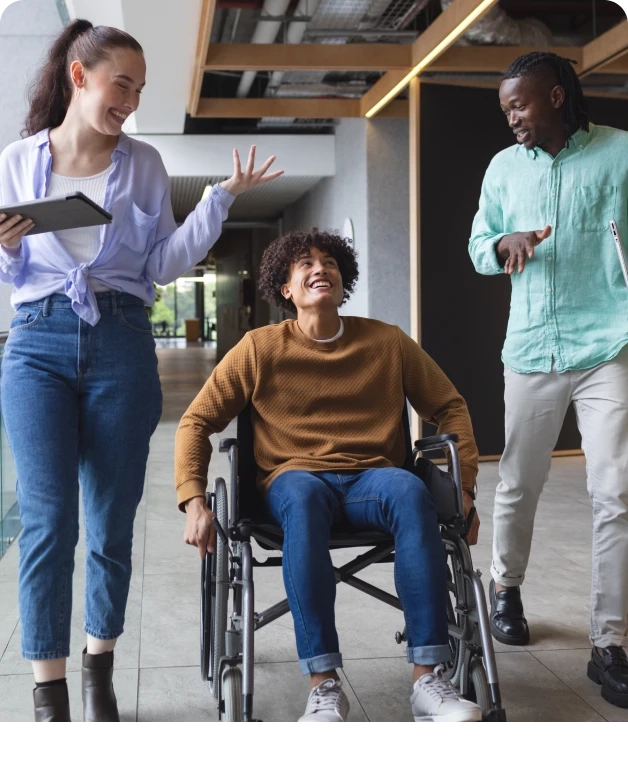 Três pessoas em um ambiente de trabalho moderno; uma delas está em uma cadeira de rodas, todas sorrindo e interagindo.